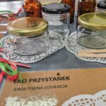 Kilka szklanych słoików i butelek z ekologicznymi kosmetykami oraz środkami czystości leży na stoliku obok kartki z napisem "Eko przystanek kreatywna edukacja".