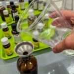Ręka wlewa płyn przez lejek do brązowej butelki, otoczonej szeregiem mniejszych pojemników z olejkami eterycznymi na zielonej macie.