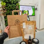 Ręka trzyma papierowy worek z ekologicznymi środkami czystości, a na stole przed nimi znajdują się słoiki z produktami i informacyjna tablica o wpływie środków czyszczących na środowisko.