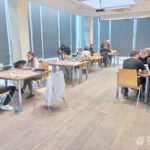 Grupa osób siedzi przy stolikach w jasnym pomieszczeniu z drewnianą podłogą i zasłoniętymi oknami.
