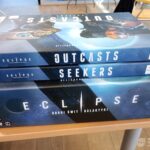 Na drewnianym stole leżą trzy pudełka gier planszowych: Outcasts, Seekers oraz Eclipse.