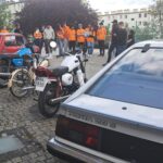Grupa osób w pomarańczowych koszulkach stoi w tle obok trzech zaparkowanych zabytkowych pojazdów: czerwonego samochodu, dwóch motocykli i srebrnego Opla Monzy 2.8 S, wszystkie na brukowanym terenie w miejskim otoczeniu.