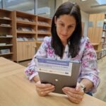 Kobieta trzyma tablet w bibliotece, prawdopodobnie uczestniczy w szkoleniu z zakresu kompetencji cyfrowych.