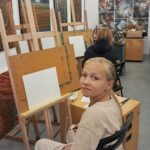 Młoda dziewczyna siedzi przed sztalugiem z pustym płótnem w pracowni malarskiej.