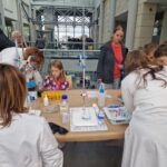 Uczniowie w białych kitach przeprowadzają doświadczenie naukowe przy stole z wyposażeniem laboratoryjnym.