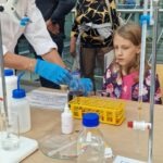 Dziewczynka obserwuje eksperyment chemiczny prowadzony przez osobę w rękawicach ochronnych.