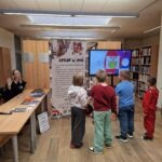 Grupa dzieci obserwuje prezentację na dużym ekranie telewizyjnym w pomieszczeniu bibliotecznym.