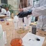 Uczniowie w rękawicach laboratoryjnych dodają ciecz do kolba podczas eksperymentu chemicznego.