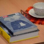 Na stole leżą dwie książki z kolorową okładką, obok filiżanka z zaparzoną kawą i okulary.