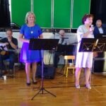 Dwóch muzyków akompaniuje dwóm wokalistkom podczas występu na scenie z nutami na stojakach.