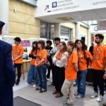 Grupa osób ubranych w identyczne pomarańczowe koszulki uważnie słucha osoby w niebieskim mundurze wewnątrz nowoczesnego budynku, w tle którego znajdują się tablice informacyjne i wyświetlacze.