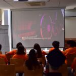 Grupa osób w pomarańczowych koszulkach siedzi w pokoju, oglądając film z angielskimi napisami na ekranie projektora. W pokoju wisi baner z napisem Miejska Biblioteka Publiczna, wskazujący na wydarzenie w bibliotece publicznej.