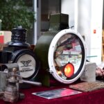 Na stole wystawowym pokrytym czerwonym materiałem znajdują się zabytkowe latarnie kolejowe, staromodne metalowe narzędzie, broszury i metalowe pudełko. Wewnątrz jednej z latarni świeci jasnopomarańczowe światło. W tle widoczna jest część osoby.