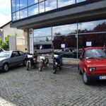 Trzy motocykle zaparkowane są między srebrnym samochodem a czerwonym kompaktowym samochodem przed szklanym budynkiem z odbiciami drzew i chmur. Chodnik jest z kostki brukowej. W tle widać furgonetkę.