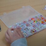 Dłonie osoby sortującej kolorowe koraliki w podzielonym plastikowym pojemniku na drewnianym stole, w pobliżu smartfon i gotowa biżuteria z koralików z boku.