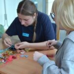 Dwie młode kobiety siedzą przy stole, skupione na nawlekaniu i tkaniu kolorowych sznurków. Jedna nosi ciemną koszulę z plakietką z imieniem; druga ma blond włosy i okulary. Na stole leżą sterty splątanych sznurków i gotowych bransoletek.
