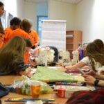 Grupa młodych ludzi siedzi i stoi wokół stołu, pracując z tkaniną i maszynami do szycia. Materiały artystyczne i kawałki tkanin są rozłożone na stole. Niektórzy noszą pasujące do siebie pomarańczowe koszule.