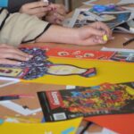 Dwie osoby pracują nad kolorowym kolażem z wyciętych obrazów, kształtów i ołówków na stole porozrzucanym skrawkami papieru i artykułami artystycznymi. Jedna osoba nosi sweter w paski.