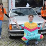 Trzech młodych ludzi pozuje przed srebrnym zabytkowym samochodem. Dwie kobiety mają na sobie pomarańczowe koszulki festiwalowe, a mężczyzna klęczy przed nimi, ubrany w kolorowy dres z różowymi, żółtymi i turkusowymi paskami.