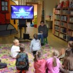 Dzieci siedzą na dywanie i słuchają osoby dorosłej, podczas gdy na ekranie wyświetlany jest film edukacyjny.