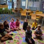 Grupa dzieci siedzi na kolorowej macie w sali lekcyjnej podczas zajęć edukacyjnych.