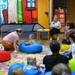 Dzieci słuchają opowiadania siedząc na dywanie w kolorowym pomieszczeniu bibliotecznym.