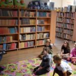 Dzieci siedzą na dywanie w bibliotece, otoczone regałami pełnymi książek podczas zajęć edukacyjnych.