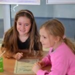 Dwie dziewczynki siedzą przy stole, przeglądając dokument i wyrażając różne emocje.