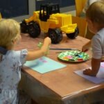 Dzieci malują farbami na stoliku, obok którego leży żółty plastikowy ładowarkę.