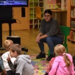 Kobieta siedzi na zielonym krześle i prowadzi zajęcia dla grupy dzieci siedzących na kolorowej macie.