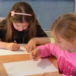 Dwie dziewczynki siedzą przy stole i piszą kredkami na białych kartkach papieru.