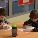 Dzieci siedzą przy stole i piszą ołówkami na papierach podczas zajęć edukacyjnych.