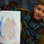 Chłopiec trzyma kartkę z rysunkiem wykonanym kredką świecową i farbami akwarelowymi, prawdopodobnie podczas eksperymentu na zajęciach.