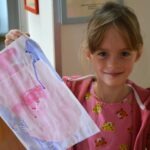 Dziewczynka trzyma w dłoniach rysunek wykonany kredkami świecowskimi i farbami akwarelowymi.