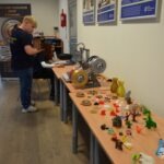 Na stole znajdują się elementy mechaniczne oraz zabawki, a w tle widoczny jest mężczyzna.