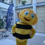Mascot pszczelego kostiumu pozuje do zdjęcia na tle budynku z flagą.