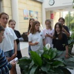 Grupa młodzieży uczestniczy w spotkaniu Dyskusyjnego Klubu Książki, skupiając się na rozmowie przy dużej donicy roślinnej.