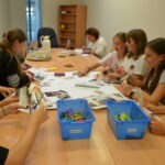 Młodzież uczestniczy w spotkaniu Dyskusyjnego Klubu Książki, pracując przy stole pełnym materiałów edukacyjnych i pojemników z klockami.