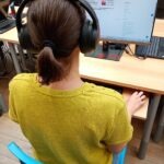 Osoba z słuchawkami, siedząca przed komputerem w bibliotece.