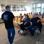 Mężczyzna wygłasza prelekcję dla grupy osób siedzących na krzesłach i w wózku inwalidzkim w jasnym pomieszczeniu.