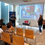 Kobieta wygłasza prezentację przed publicznością siedzącą na krzesłach, a na ekranie wyświetlana jest ilustracja związana z bezpieczeństwem cyfrowym.