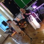 Dwóch muzyków z zespołu śpiewa do mikrofonów na scenie podczas koncertu w sali gimnastycznej.