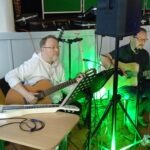 Dwóch muzyków siedzi na krzesłach i gra na gitarach podczas występu zespołu.