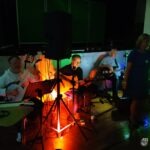 Muzycy zespołu grają na instrumentach w słabo oświetlonym pomieszczeniu, otoczeni sprzętem nagłaśniającym.