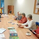 Seniorzy siedzą przy długim stole, przeglądając książki i notatki podczas spotkania poświęconego pielęgnacji roślin.