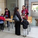 Kobieta w stroju wolontariusza rozmawia z kobietą i dwoma dziećmi przy stoliku podczas wydarzenia w bibliotece.