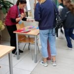 Kobieta wręcza uczestnikowi wydarzenia materiały informacyjne przy stoliku z prezentacjami.