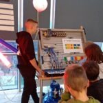 Mężczyzna instruuje dzieci przy stanowisku z panelem edukacyjnym wyposażonym w elementy robotyki.
