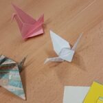 Na drewnianym stole leżą cztery papierowe żurawie w różnych kolorach: różowy, biały, zielono-niebieski oraz żółty.