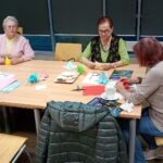 Seniorzy siedzą przy stole i pracują z papierem kolorowym oraz narzędziami do rękodzieła.